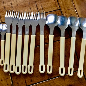 Op de afbeelding: Een set van 18 zilverkleurige vorken en lepels met ivoorwitte handvatten. De handvatten hebben een eenvoudig, afgerond ontwerp met een klein gaatje aan het einde.