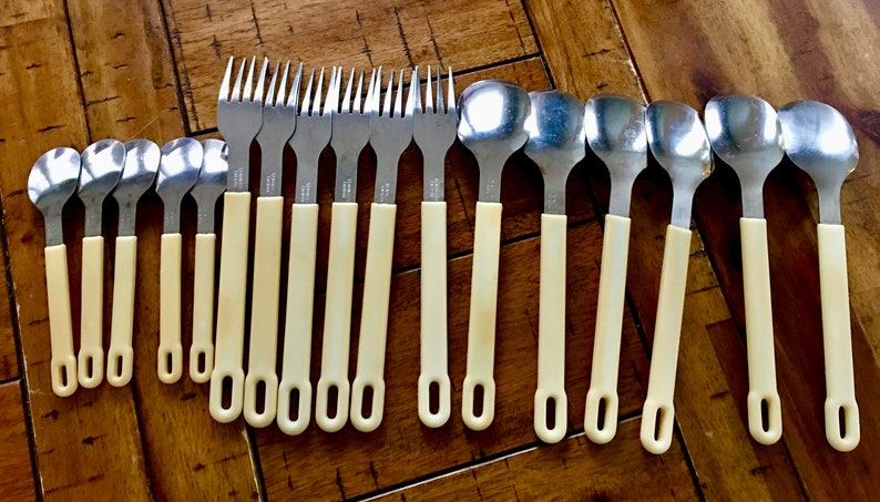 Op de afbeelding: Een set van 18 zilverkleurige vorken en lepels met ivoorwitte handvatten. De handvatten hebben een eenvoudig, afgerond ontwerp met een klein gaatje aan het einde.