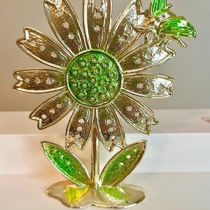 May include: A gold-colored metal flower sculpture with green accents. The flower has detailed petals with small holes, a green center, and green leaves. A green bee is perched on the flower.