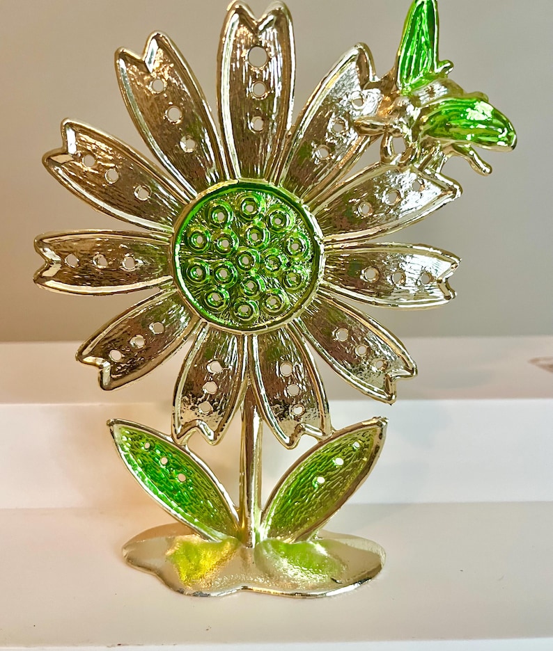 May include: A gold-colored metal flower sculpture with green accents. The flower has detailed petals with small holes, a green center, and green leaves. A green bee is perched on the flower.