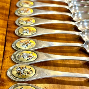 May include: A set of twelve silver spoons with gold-toned handles. Each handle features a different animal illustration.