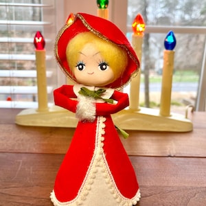 May include: A vintage Christmas doll with a red velvet dress and hood, trimmed with white and gold accents. The doll has a blonde head and a sweet, painted face. The doll is holding its arms in front of itself. The background includes a candle holder with colorful lights.