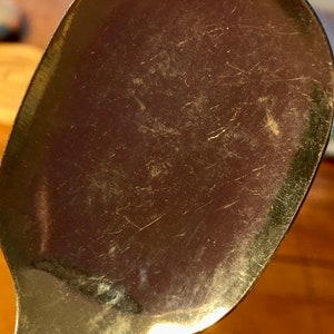 Op de afbeelding: Een close-up van een zilveren lepel met een glanzend oppervlak en zichtbare krassen. De lepel is licht gebogen en heeft een afgeronde kom.