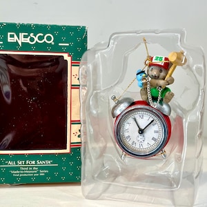 May include: A Christmas-themed ornament featuring a red alarm clock with a bear figurine holding a mallet. The bear wears a green shirt and a hat with the number 25. The ornament is in a clear plastic case with a green box that reads "ALL SET FOR SANTA".