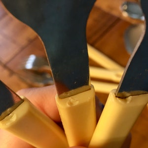 Op de afbeelding: Close-up van drie zilveren lepels met cr&egrave;mekleurige kunststof handvatten. De lepels worden in een hand gehouden, en de handvatten zijn zichtbaar. De lepels zijn allemaal even groot en even gevormd.