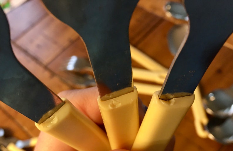 Op de afbeelding: Close-up van drie zilveren lepels met cr&egrave;mekleurige kunststof handvatten. De lepels worden in een hand gehouden, en de handvatten zijn zichtbaar. De lepels zijn allemaal even groot en even gevormd.
