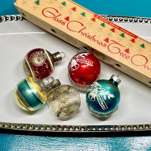 Op de afbeelding: Een verzameling vintage glazen kerstboomversieringen in verschillende kleuren, waaronder rood, groen en helder. Sommige ornamenten hebben strepen of bloemmotieven, en één heeft het woord "Greetings". De ornamenten worden gepresenteerd op een wit dienblad met een decoratieve rand, naast een vintage doos.