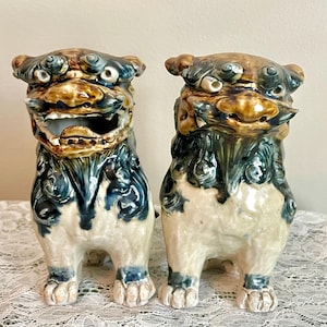 May include: Two ceramic guardian lion statues. The statues are in a seated position, with detailed features including open mouths, and intricate mane and body details. The color palette includes shades of brown, blue, and cream, giving them an antique appearance.