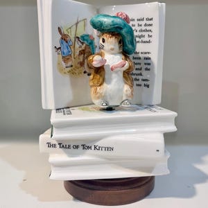 May include: A ceramic figurine of a rabbit wearing a teal beret and brown jacket, standing on a stack of white books. The top book is open, displaying an illustration and text. The bottom book is titled The Tale of Tom Kitten. The base is wood.