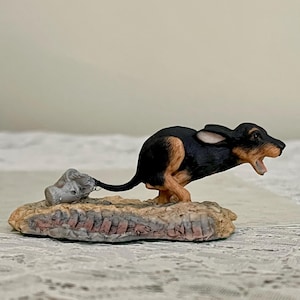 May include: A black and brown figurine of a bilby, a small marsupial native to Australia, running with its mouth open. The figurine is on a rocky base with a small, gray, textured object behind it.