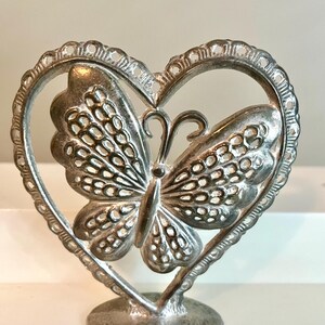 May include: A silver-colored butterfly figurine within a heart-shaped frame. The butterfly has detailed wing patterns and is mounted on a round base. The heart frame has a decorative, scalloped edge.