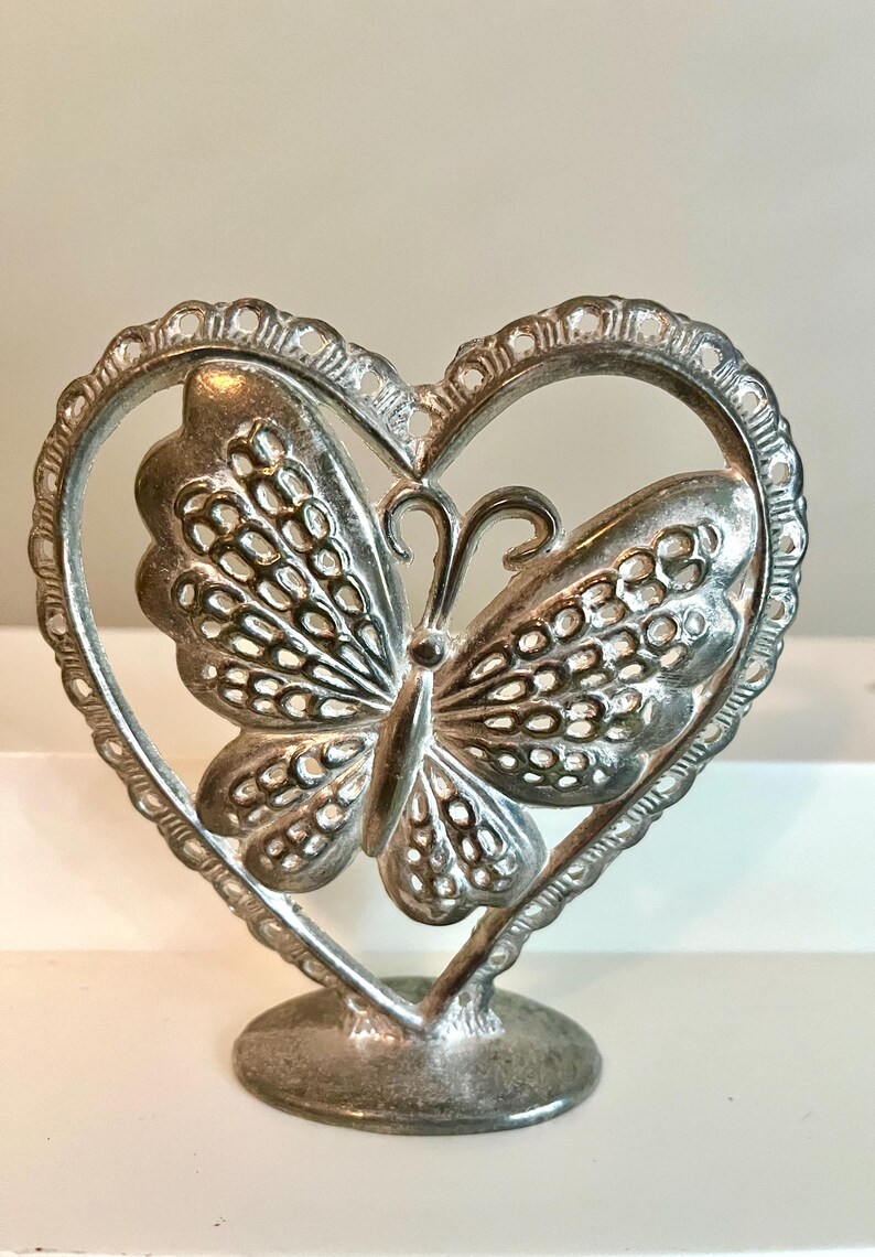 May include: A silver-colored butterfly figurine within a heart-shaped frame. The butterfly has detailed wing patterns and is mounted on a round base. The heart frame has a decorative, scalloped edge.