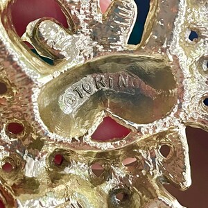 May include: Close-up of a gold-toned metal brooch with the word "TORINO" stamped on the surface. The brooch has an ornate design with openwork and red accents.