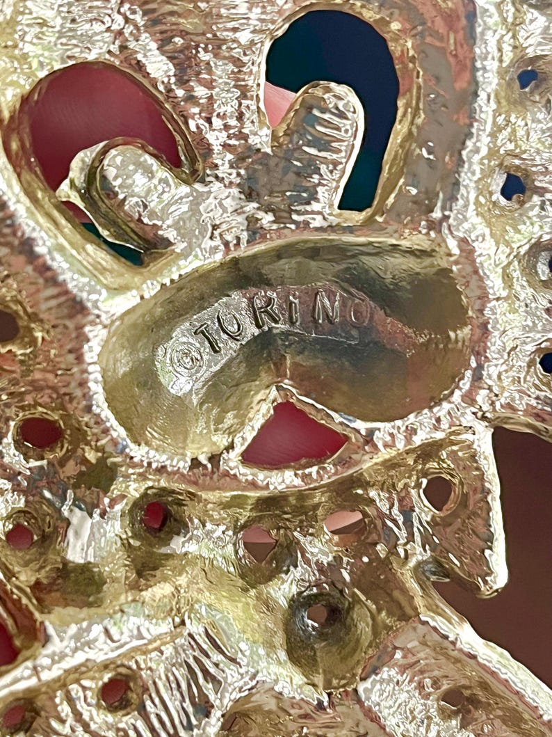 May include: Close-up of a gold-toned metal brooch with the word "TORINO" stamped on the surface. The brooch has an ornate design with openwork and red accents.