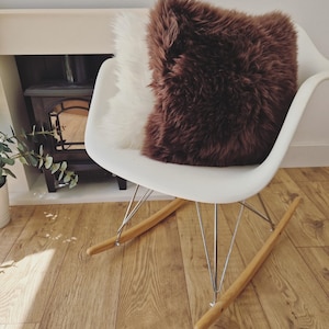 May include: A white rocking chair with a brown and white fluffy pillow. The chair has a white seat, metal legs, and wooden rockers. The pillows have a shaggy texture, adding a cozy element to the room.