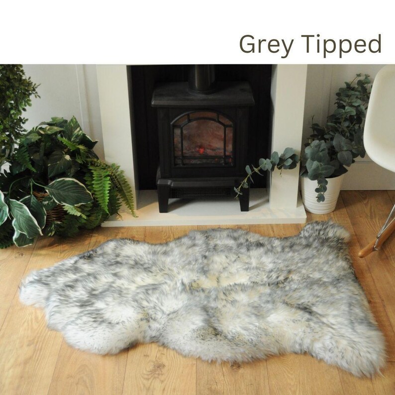 May include: A white faux fur rug with grey tips. The rug is laid out on a wooden floor in front of a fireplace.