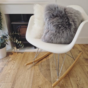 May include: A white rocking chair with a gray and white fluffy pillow. The chair has wooden rockers and metal legs. The background includes a fireplace and hardwood flooring.