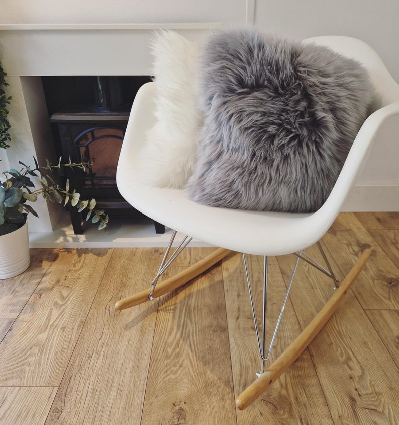 May include: A white rocking chair with a gray and white fluffy pillow. The chair has wooden rockers and metal legs. The background includes a fireplace and hardwood flooring.