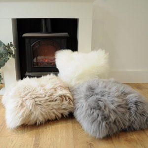 May include: Three fluffy, faux fur cushions in cream, tan, and gray hues rest on a wooden floor. The cushions have a rectangular shape and a shaggy texture. A white fireplace and a decorative plant are in the background.