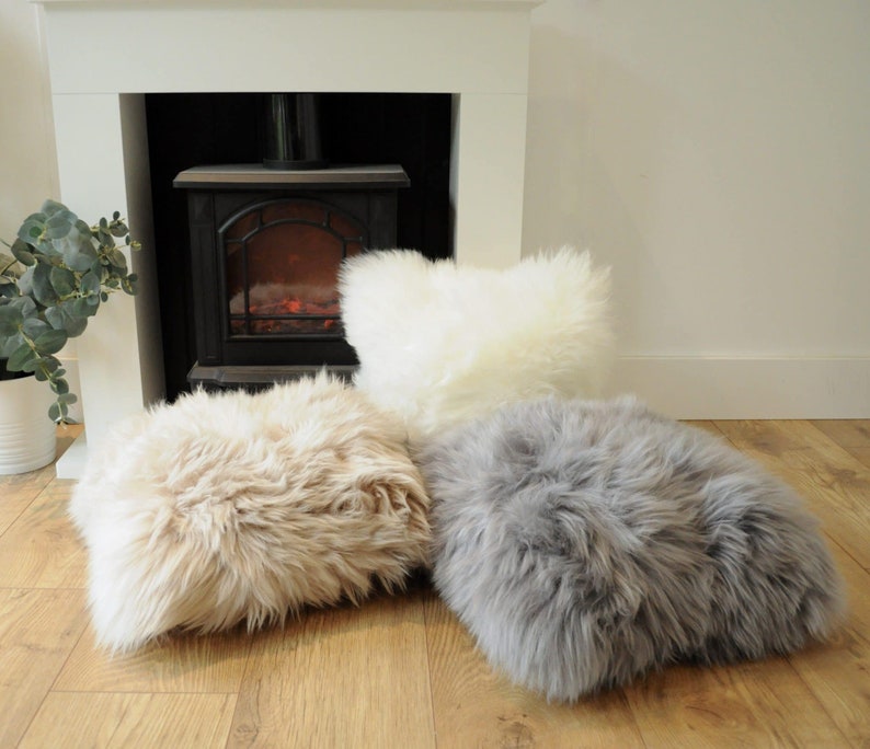 May include: Three fluffy, faux fur cushions in cream, tan, and gray hues rest on a wooden floor. The cushions have a rectangular shape and a shaggy texture. A white fireplace and a decorative plant are in the background.