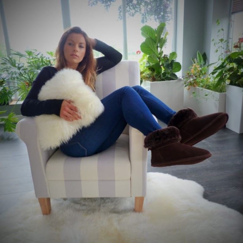 May include: A white fluffy pillow is held by a person sitting in a striped armchair. The person wears blue jeans and brown suede boots with fur trim. The chair sits on a white sheepskin rug.