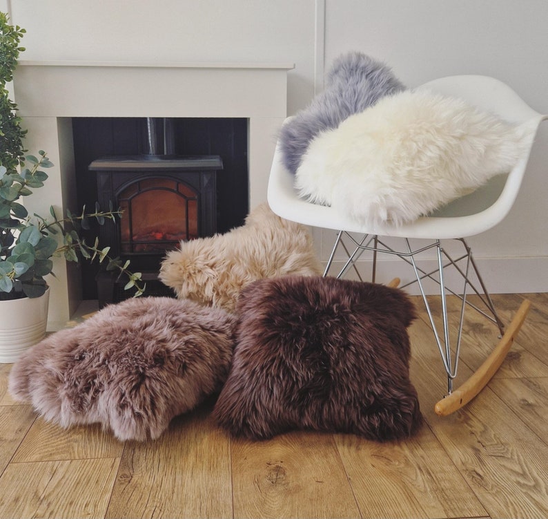 May include: A collection of fluffy, textured throw pillows in various colors: cream, gray, tan, and brown. The pillows are arranged on a wooden floor, with some resting on a white chair. A fireplace and greenery are visible in the background.