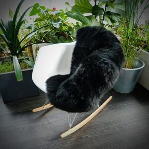 May include: A black sheepskin rug placed on a white rocking chair with wooden arms. The chair is in a room with plants in the background.