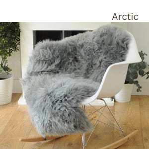 May include: A gray sheepskin rug is draped over a white rocking chair. The rug is soft and fluffy, and the chair is made of metal and wood. The rug is labeled "Arctic".