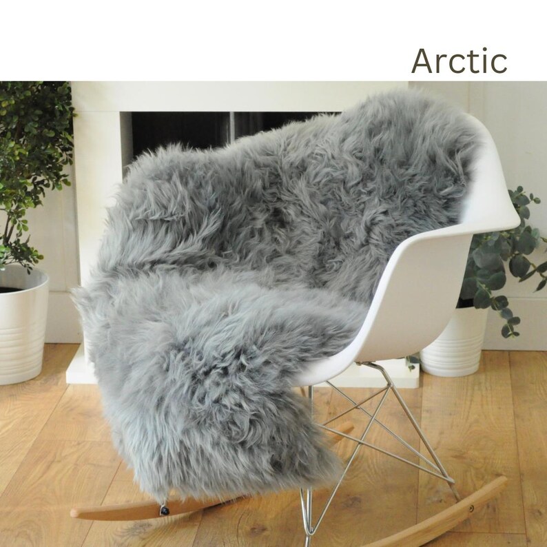 May include: A gray sheepskin rug is draped over a white rocking chair. The rug is soft and fluffy, and the chair is made of metal and wood. The rug is labeled "Arctic".