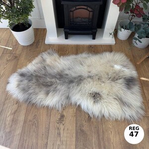 May include: A rectangular sheepskin rug in shades of white, grey, and brown. The rug is made of long, soft fibres and lies on a wooden floor. The rug has a natural, textured appearance, and is a decorative item.