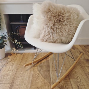 May include: A white rocking chair with a fluffy, beige pillow. The chair has wooden rockers and metal legs. The pillow is a light brown color and has a shaggy texture. The chair is on a wooden floor.