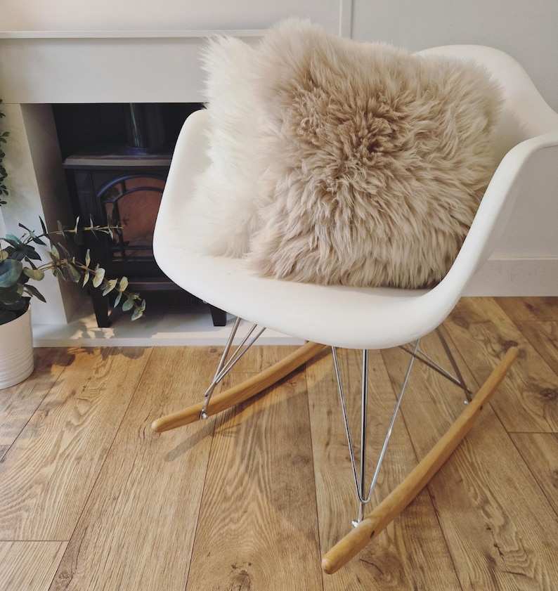 May include: A white rocking chair with a fluffy, beige pillow. The chair has wooden rockers and metal legs. The pillow is a light brown color and has a shaggy texture. The chair is on a wooden floor.