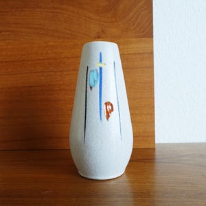 Könnte beinhalten: Eine weiße Keramikvase mit strukturierter Oberfläche. Die Vase hat ein Design aus vertikalen schwarzen und blauen Linien mit abstrakten blauen, gelben und roten Formen. Die Vase hat einen abgerundeten Boden und verjüngt sich nach oben.