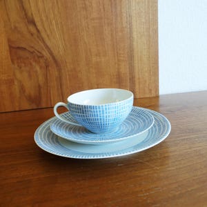 May include: A blue and white patterned teacup and saucer set. The cup and saucers have a grid pattern design. The set includes a teacup, a small plate, and a larger plate. The set is on a wooden surface.