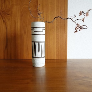 May include: A white ceramic vase with a cylindrical shape, adorned with dark gray geometric patterns. The vase is decorated with horizontal bands and vertical lines. A dried branch with small, brown buds extends from the vase. The vase is on a wooden surface.