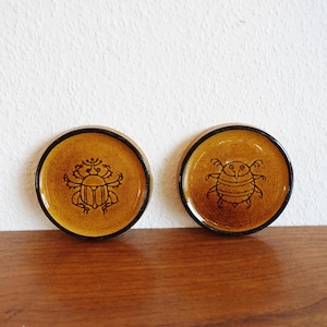 May include: Two brown ceramic coasters with black outlines. Each coaster features a different stylized beetle design.