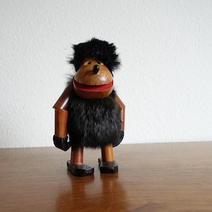 May include: A wooden toy figure with a black fur coat, red mouth, and brown wooden arms and legs. The toy has a black fur hat and is standing on a wooden surface.