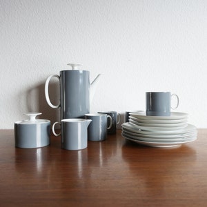 Op de afbeelding: Een grijs en wit keramisch theeservies met een theepot, roomkan, suikerpot, vier kopjes en zes schoteltjes. De set is op een houten tafel gerangschikt.