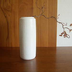 Op de afbeelding: Een hoge, witte keramische vaas met een gestructureerd, gestippeld patroon. De vaas is cilindrisch en staat op een donker houten oppervlak. Een droge, takkerige tak strekt zich uit vanaf de bovenkant van de vaas.
