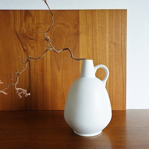 May include: A white ceramic vase with a rounded body and a handle. A dried, brown branch extends from the vase. The vase is set against a wooden background.