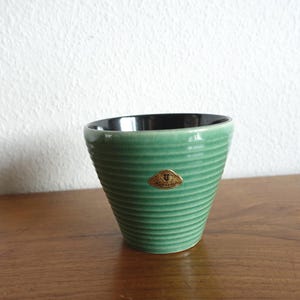 Könnte beinhalten: Eine grüne Keramiktasse mit schwarzem Innenraum. Die Tasse hat ein geripptes Design und ein kleines goldfarbenes Etikett. Die Tasse steht auf einer Holzoberfläche.