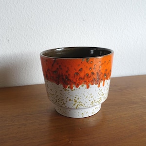 Könnte beinhalten: Ein Keramik-Pflanztopf mit einem unverwechselbaren Design. Die obere Hälfte ist leuchtend orange, geht in eine weiße Basis über, die mit Grün und Braun gesprenkelt ist. Der Innenraum ist dunkelbraun. Der Topf hat einen abgestuften Sockel.