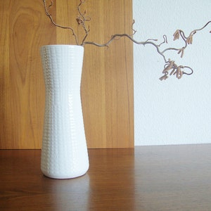 May include: A white ceramic vase with a textured surface, featuring a slender, cylindrical shape. The vase is empty and sits on a wooden surface. A dried branch with brown twigs extends from the vase.