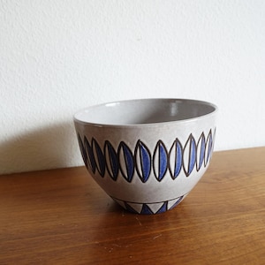 Könnte beinhalten: Eine graue Keramikschale mit einem blau-schwarzen geometrischen Muster. Die Schale hat eine abgerundete Form und eine weite Öffnung. Das Design zeigt blaue Ovale, die mit Schwarz umrandet sind. Die Schale steht auf einer Holzoberfläche.