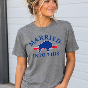 Könnte beinhalten: Ein graues, kurzärmeliges T-Shirt mit dem blauen Schriftzug MARRIED INTO THIS und einer blauen Büffelsilhouette, die über einem horizontalen roten Streifen zentriert ist. Die Grafik ist auf der Vorderseite des Rundhals-Shirts aus Baumwollmischgewebe.