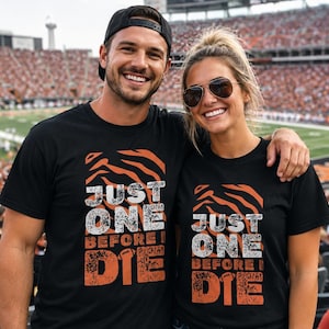 May include: Two black short-sleeved t-shirts featuring a graphic design with orange tiger stripes at the top and the text "JUST ONE BEFORE I DIE" in white and orange, with a small football icon replacing the letter "I" in the word "DIE".