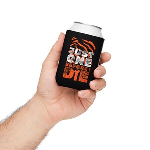 May include: A black foam can cooler featuring an orange tiger stripe pattern at the top and the white and orange distressed text "JUST ONE BEFORE I DIE" with a small football icon replacing the letter O in the word ONE.