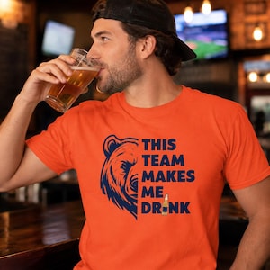 May include: A bright orange short-sleeved t-shirt featuring a dark blue graphic of a bear's face on the left side and the text "THIS TEAM MAKES ME DRINK" on the right, with a small illustration of a beer bottle replacing the letter I.