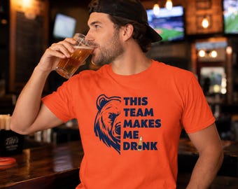 May include: A bright orange short-sleeved t-shirt featuring a dark blue graphic of a bear's face on the left side and the text "THIS TEAM MAKES ME DRINK" on the right, with a small illustration of a beer bottle replacing the letter I.