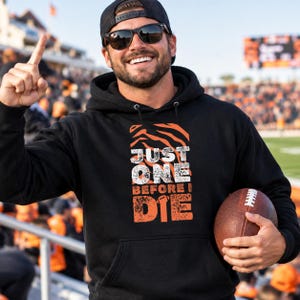 Op de afbeelding: Een zwarte hoodie met een grafische print met oranje tijgerstrepen aan de bovenkant en de witte en oranje tekst "JUST ONE BEFORE I DIE" met een klein football-icoontje dat de letter I in het woord DIE vervangt, en een voorzak.
